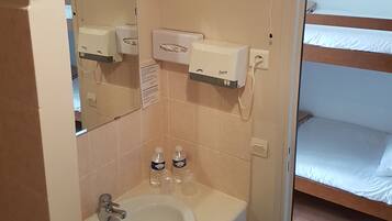 A bathroom with a sink, mirror, hand dryer, and a heater. There is a bed with a wooden headboard and white bedding.