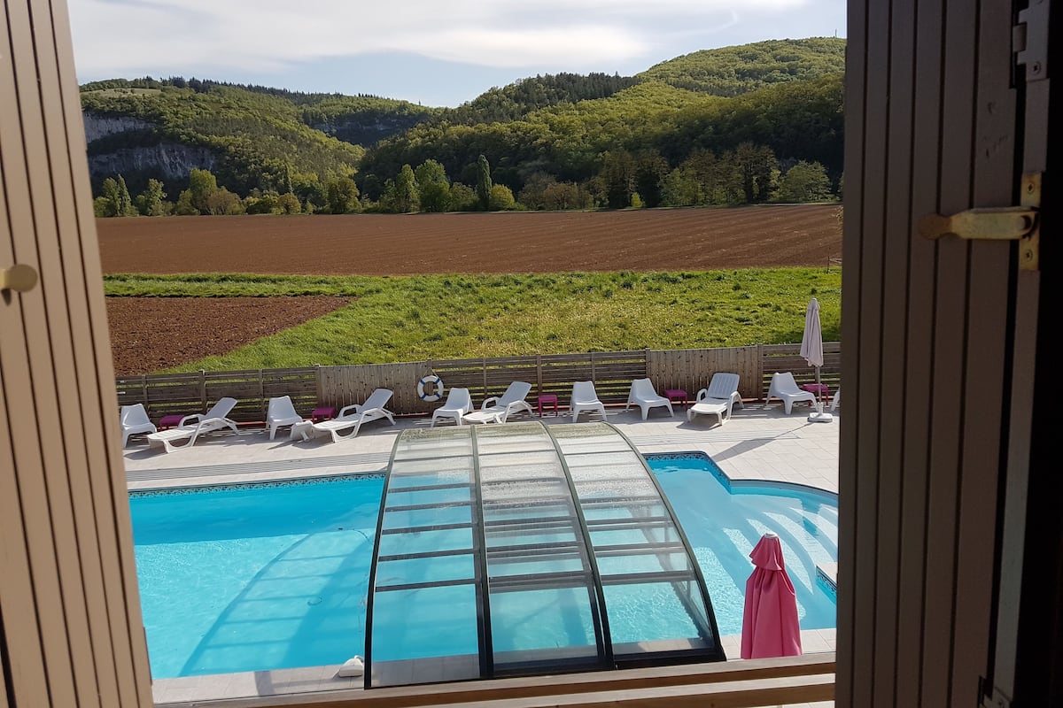 Zona de piscina con tumbonas y una sección con paredes de vidrio, con vistas a un paisaje pintoresco con colinas y campos.
