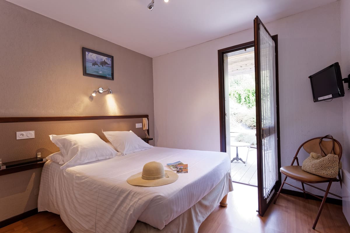 Una cama bien hecha con sábanas blancas, un sombrero y un bolso sobre una silla de madera, un televisor en la pared, y una puerta que da al exterior.