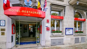 Entrada de un hotel con un llamativo letrero rojo que dice "HOTEL-RESTAURANT" y banderas de Francia, Bélgica y el Reino Unido.