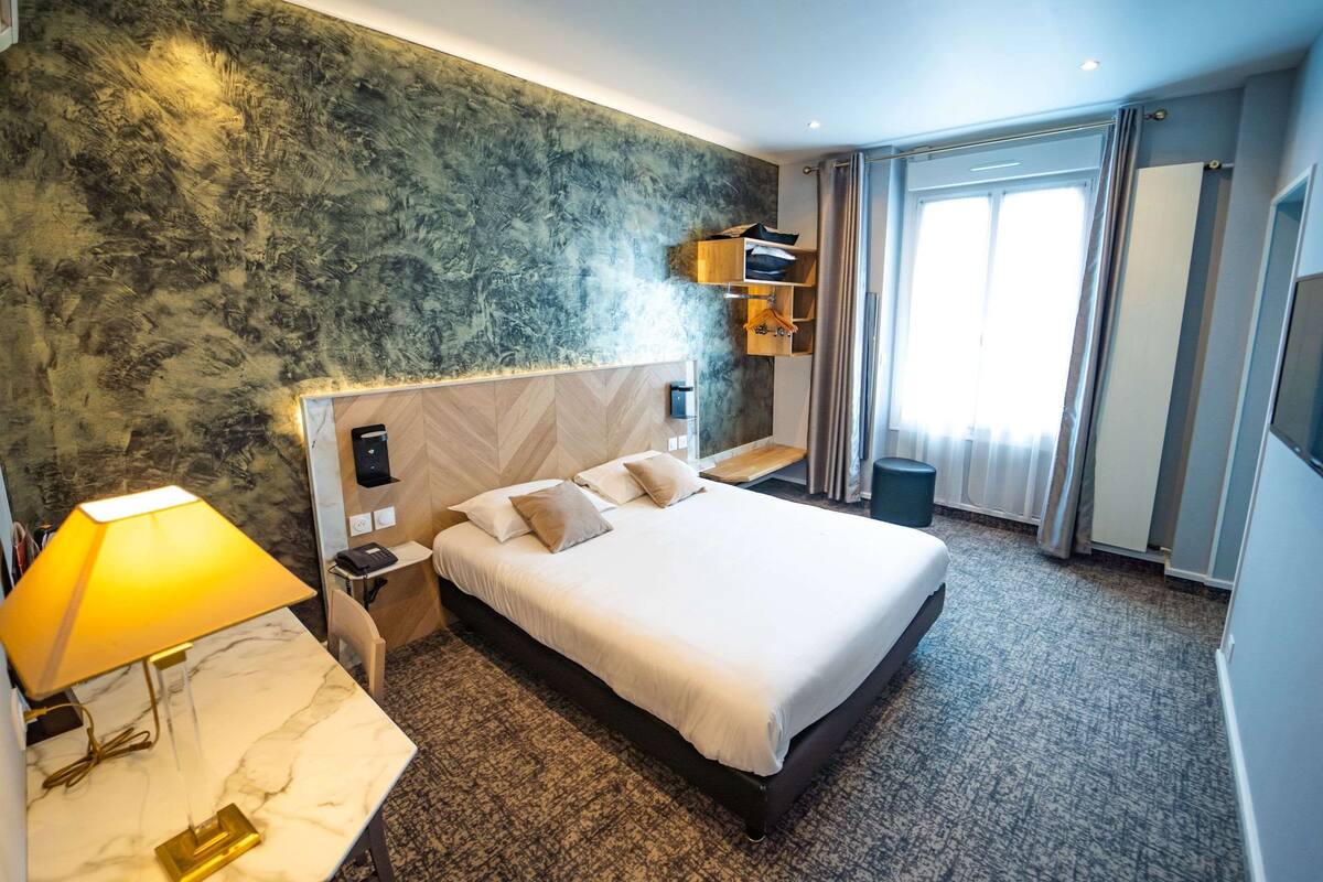 A hotel room with a large bed, a nightstand, a lamp, a wooden headboard, a marble table, a wall-mounted shelf, and a window with curtains.