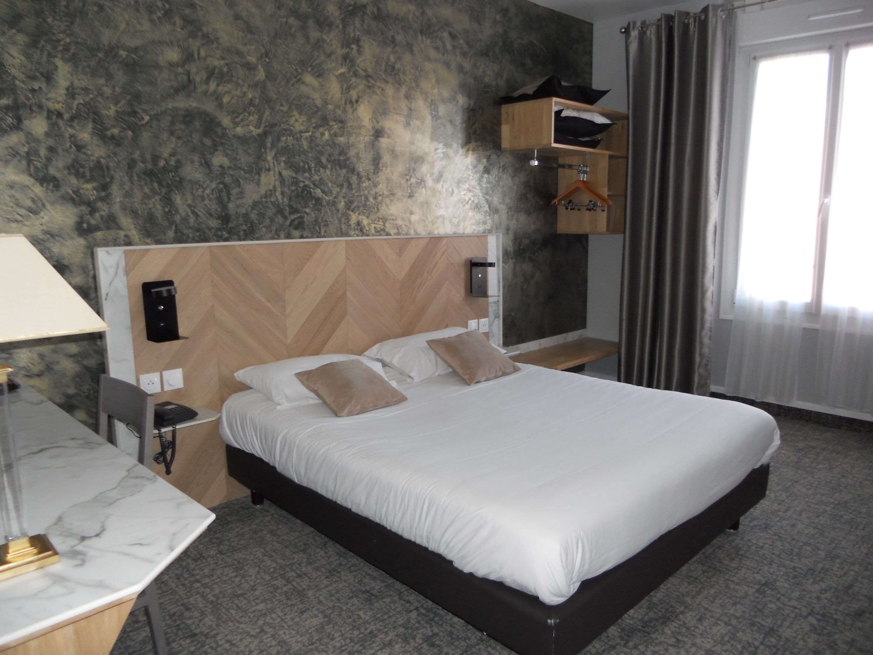 Habitación de hotel con una cama grande, una mesita de noche, una lámpara, cabecero de madera, estante en la pared y una ventana con cortinas.