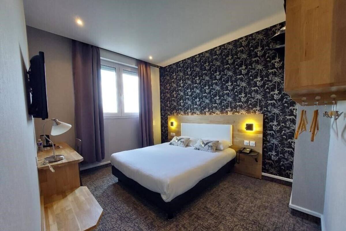 A hotel room with a bed, bedside table, lamp, and a wall with a floral pattern.