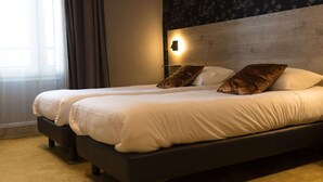 Cama doble con ropa blanca y dos almohadas marrones, una mesita de noche y una lámpara fijada a la pared.