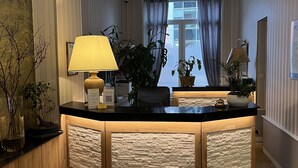 Zona de recepción con mostrador de madera, lámpara colgante, plantas en macetas y vista a través de una ventana con cortinas.