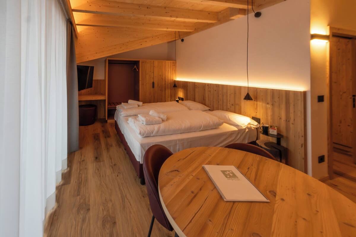 A cozy room with a wooden ceiling, a bed with white linens, a round wooden table, and a small desk with a lamp.
