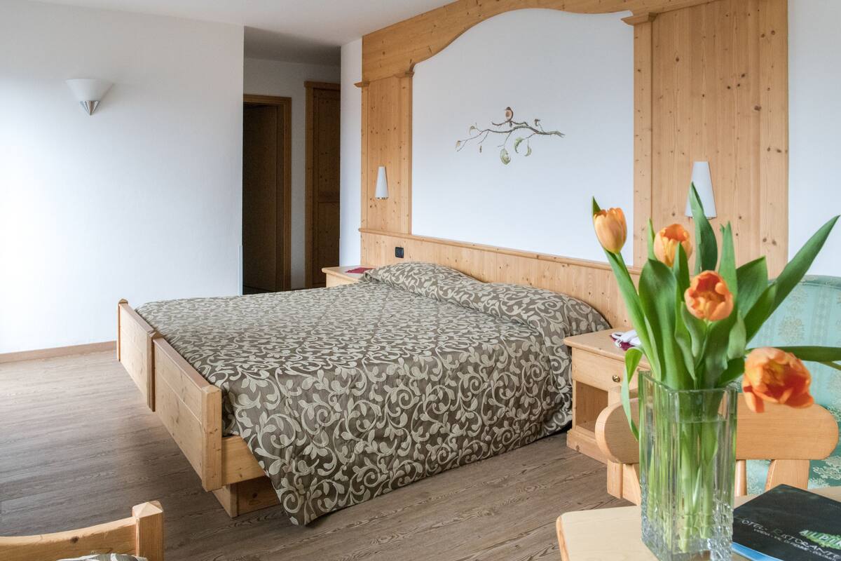 A bedroom with a wooden bed, a nightstand, a vase with flowers, and a wall-mounted lamp.