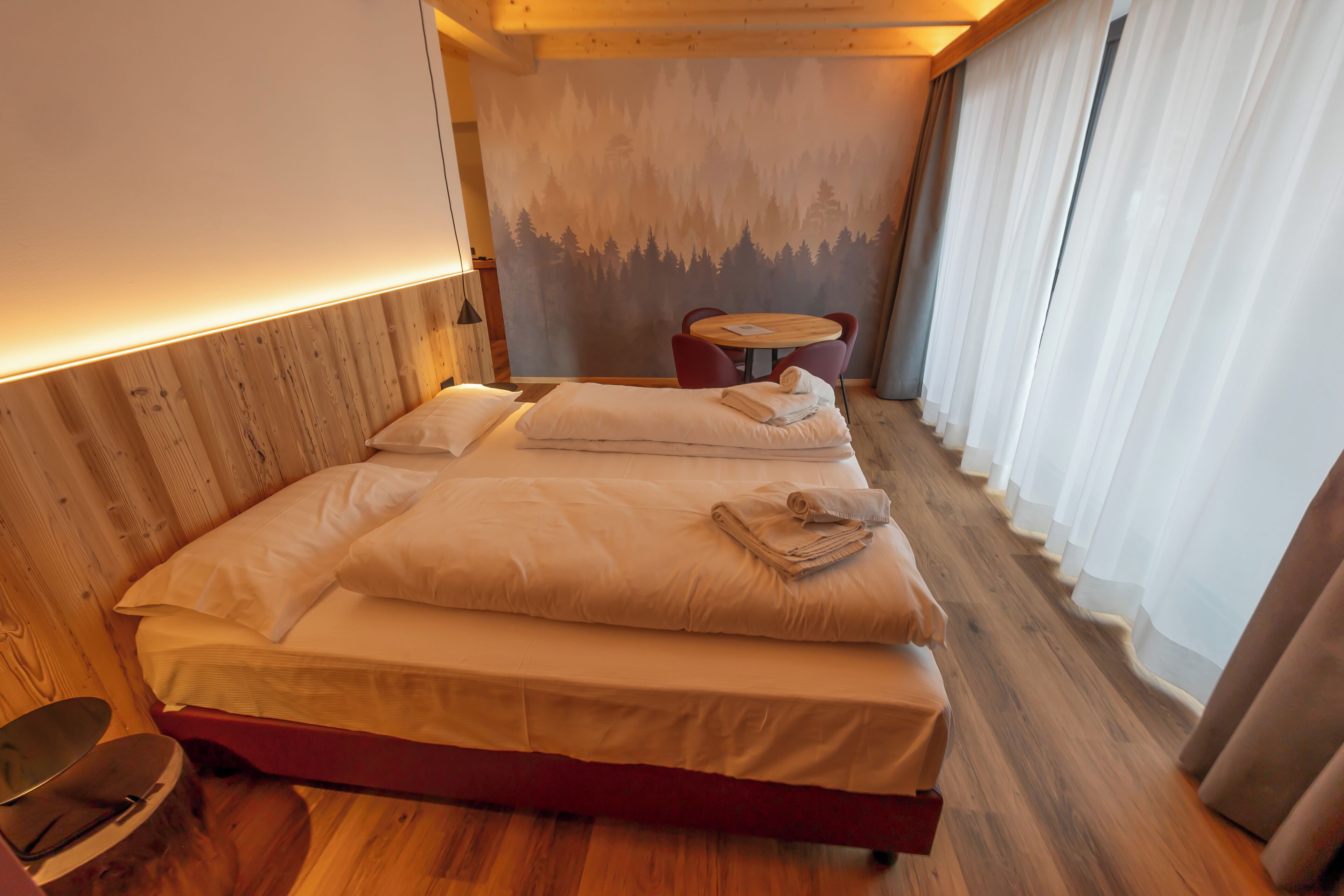 A cozy room with a wooden headboard, a bed with neatly folded towels, a small round table, and a view of a forest through sheer curtains.