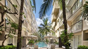 A poolside area with lounge chairs and umbrellas, surrounded by palm trees and a building with balconies. - The Akmani Legian (Kuta)