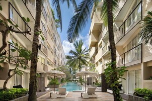 A poolside area with lounge chairs and umbrellas, surrounded by palm trees and a building with balconies.