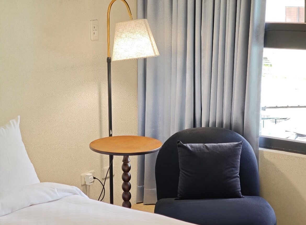A hotel room with a bed, a chair, a small table, and a lamp.