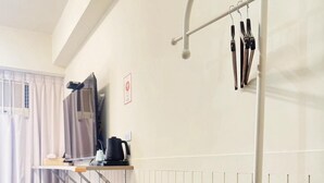A room with a wooden floor, a small round table, a white wall-mounted shelf with a kettle, a TV, and a pair of scissors.