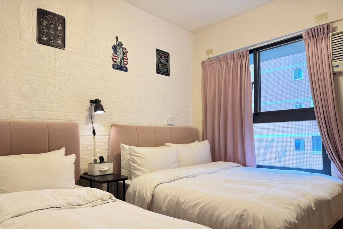 A hotel room with two beds, a nightstand with a lamp, and a window with curtains.