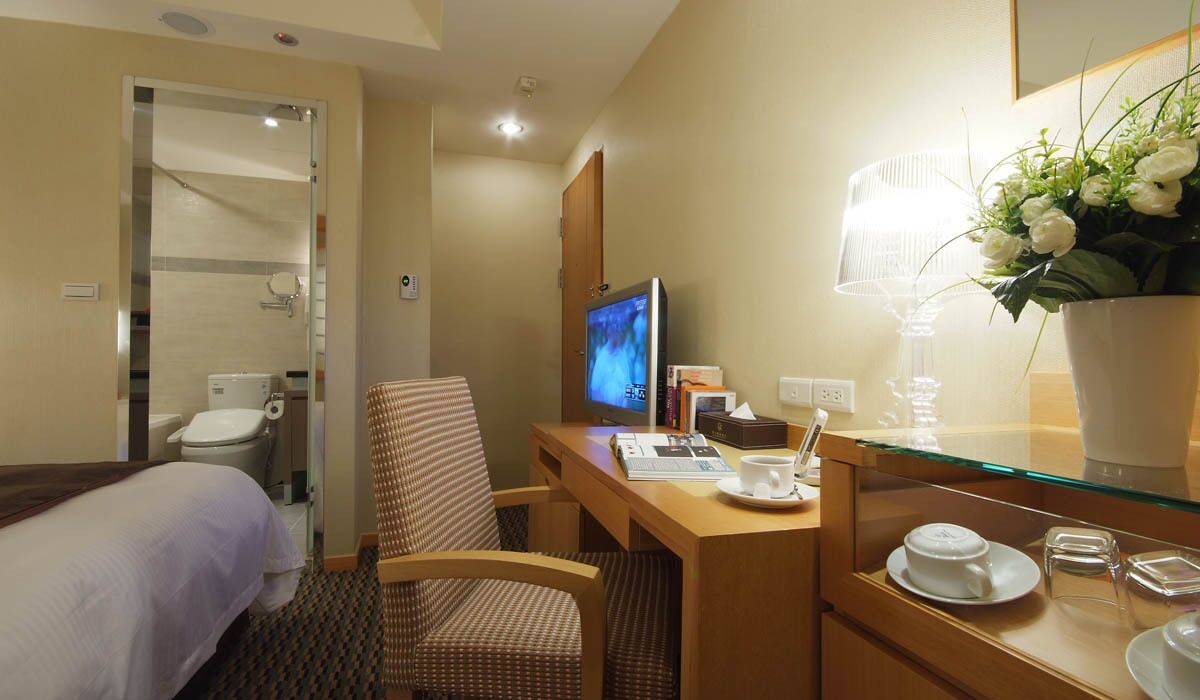 A hotel room with a bed, a desk with a computer, a chair, a desk lamp, a vase with flowers, and a bedside table with a cup and saucer.