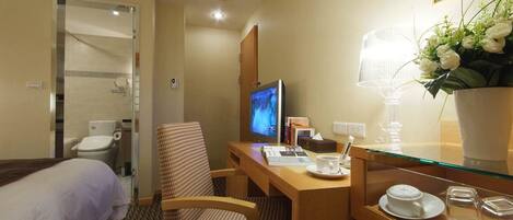 A hotel room with a bed, a desk with a computer, a chair, a desk lamp, a vase with flowers, and a bedside table with a cup and saucer.