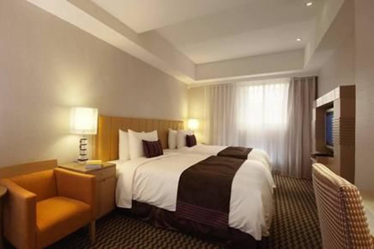 A hotel room with a large bed, a nightstand, a lamp, a chair, and a view of the outdoors through the window.