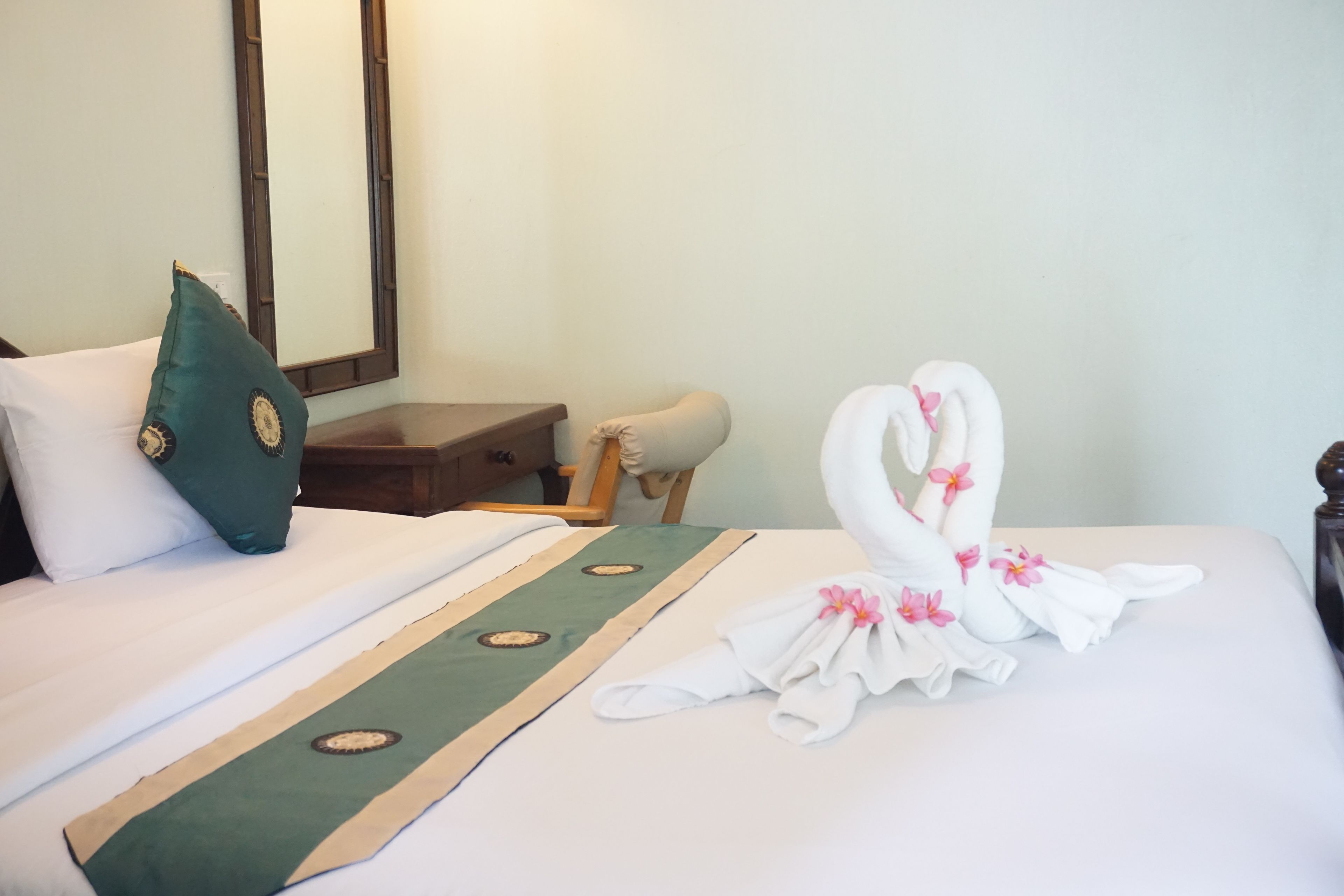 A hotel room with a neatly made bed, a green throw pillow, a wooden nightstand, and a mirror on the wall.