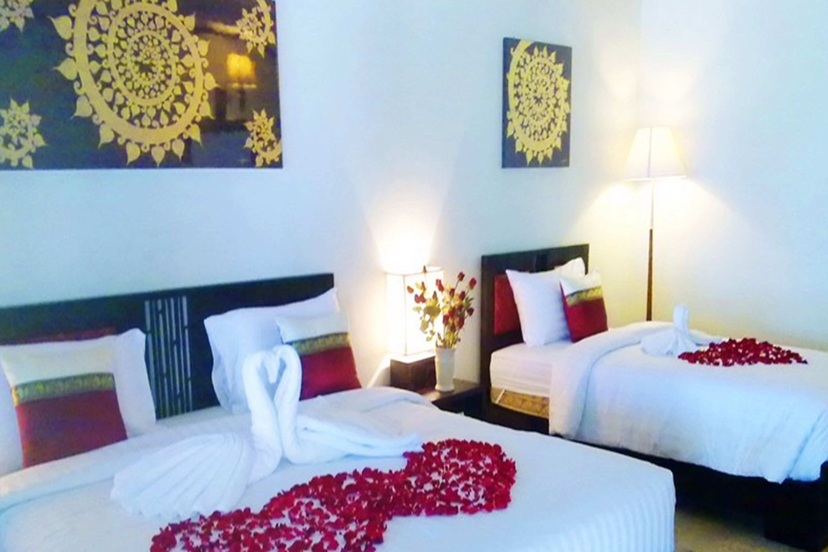 Een hotelkamer met twee bedden, elk voorzien van witte linnens en rode rozenblaadjes. Op de nachtkastjes staan kunstwerken en een lamp.