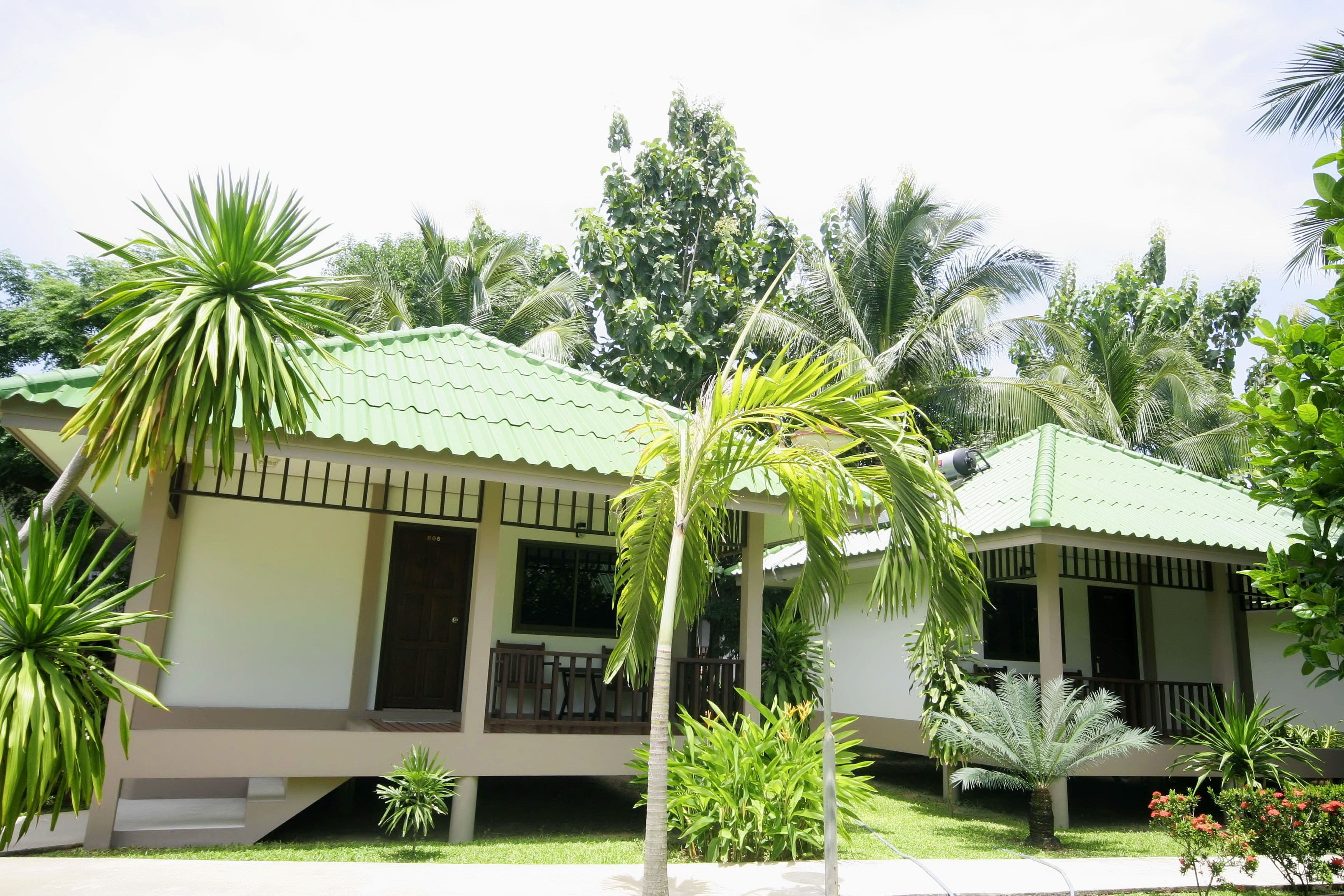 A two-story building with a green roof, surrounded by lush greenery including palm trees and other tropical plants.