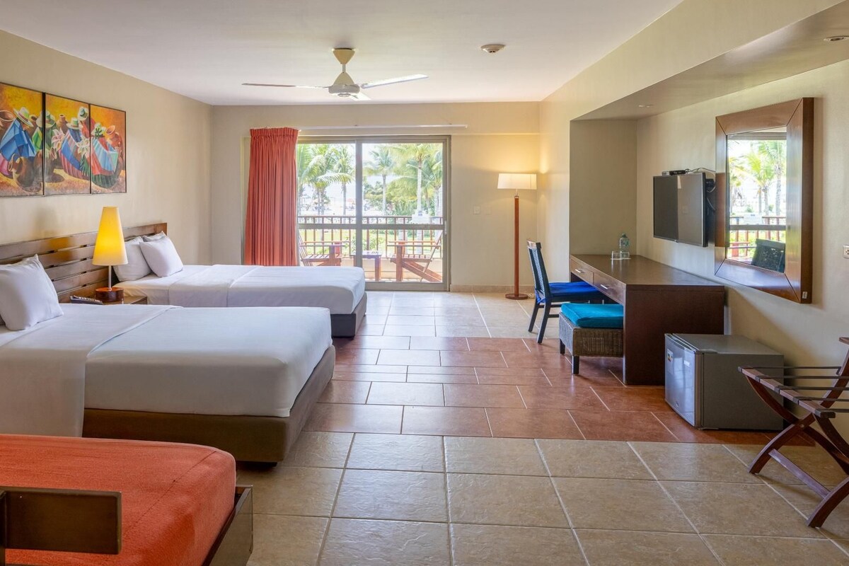 Habitación de hotel con dos camas, un escritorio, una silla, un televisor y un balcón con vistas a palmeras.