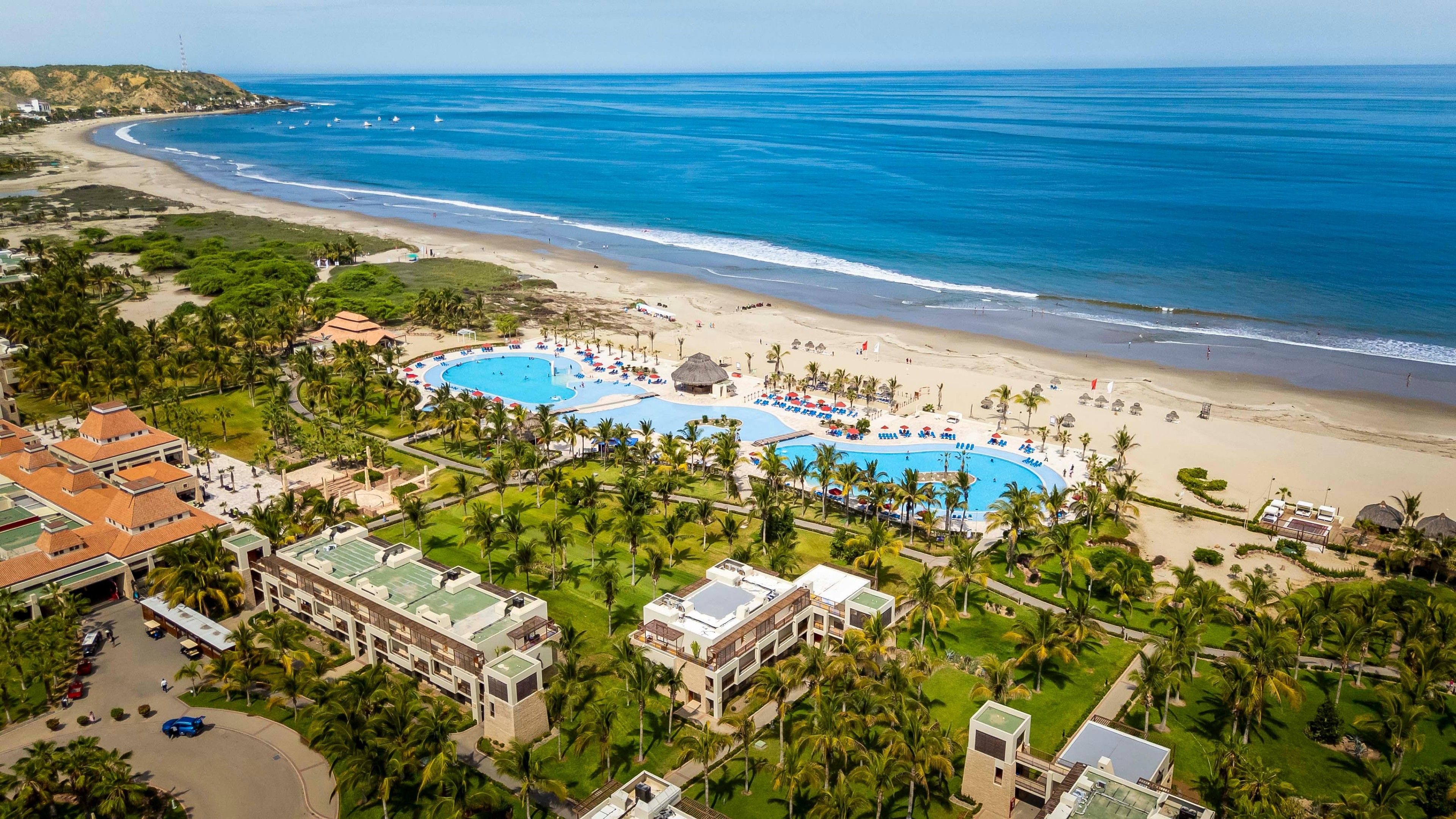 Un complex turístic amb una àrea de piscina àmplia, davant de la platja i vegetació exuberant.