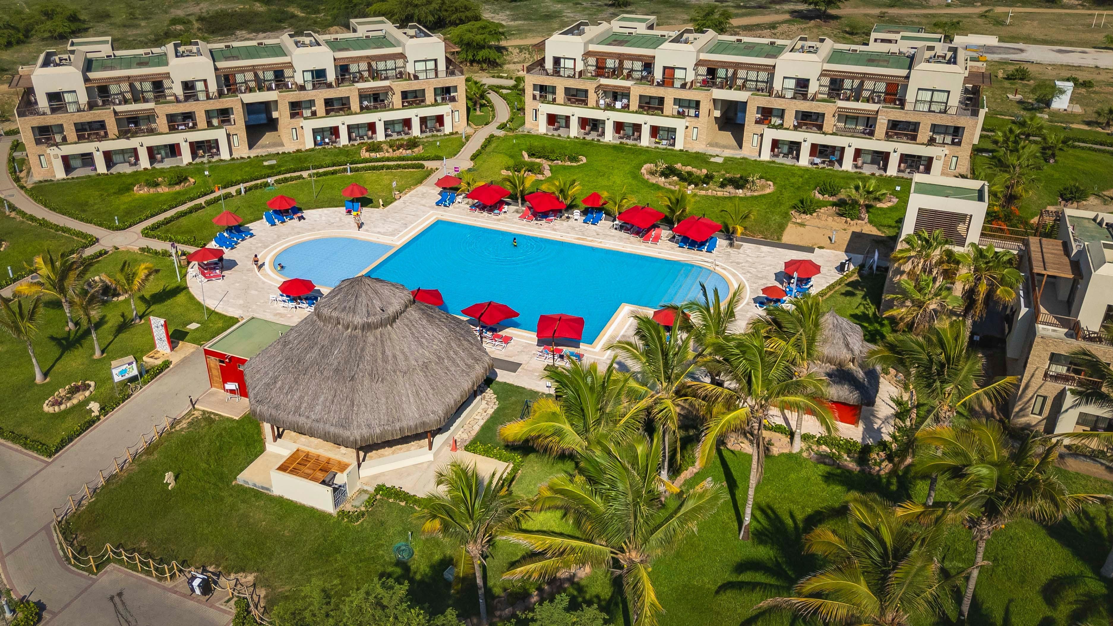 Un resort con varias construcciones, una piscina central y palmeras aledañas.
