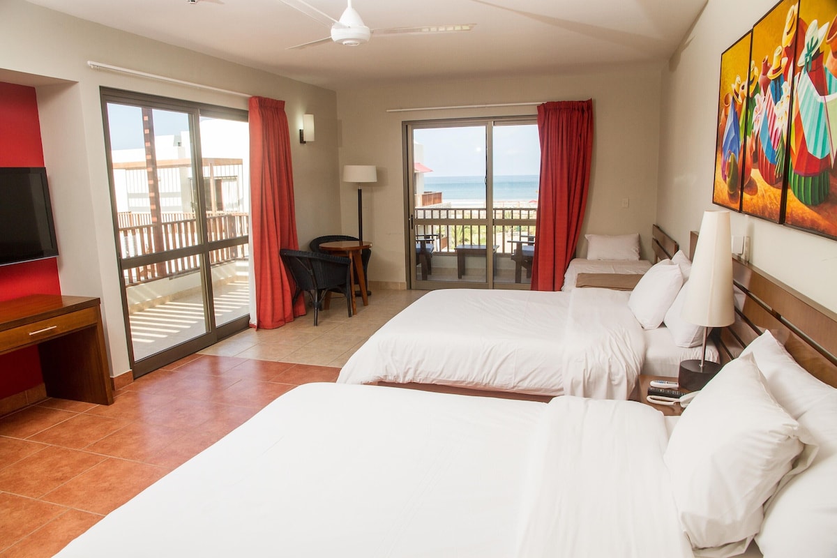 A hotel room with two beds, a desk, a chair, a TV, and a balcony with a view of the sea.