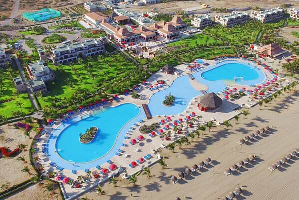 A resort with two large swimming pools, a beach area with sun loungers, and a well-maintained lawn.
