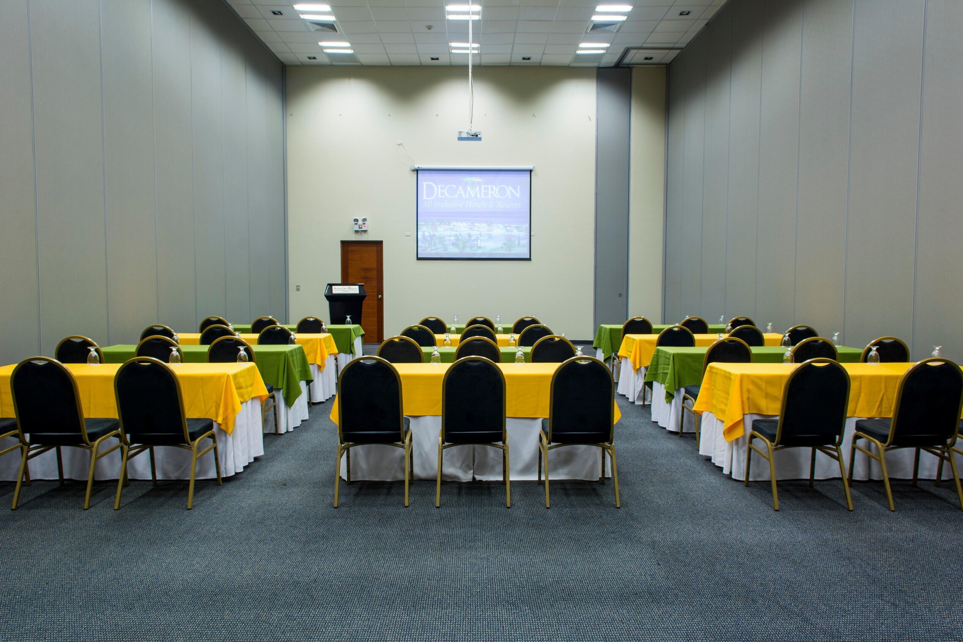 Un salón de conferencias con mesas y sillas dispuestas en filas, una pantalla de proyector y una puerta.