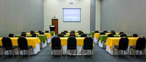 Un salón de conferencias con mesas y sillas dispuestas en filas, una pantalla de proyector y una puerta.