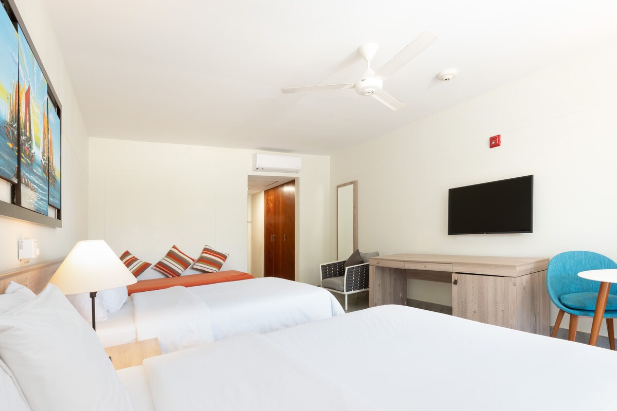 A hotel room with two beds, a desk, a chair, a TV, and a ceiling fan.