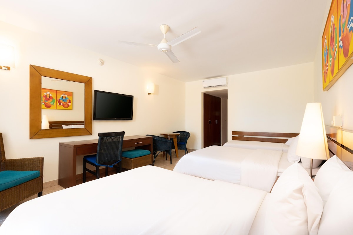 A hotel room with two beds, a desk with chairs, a television, and a ceiling fan.