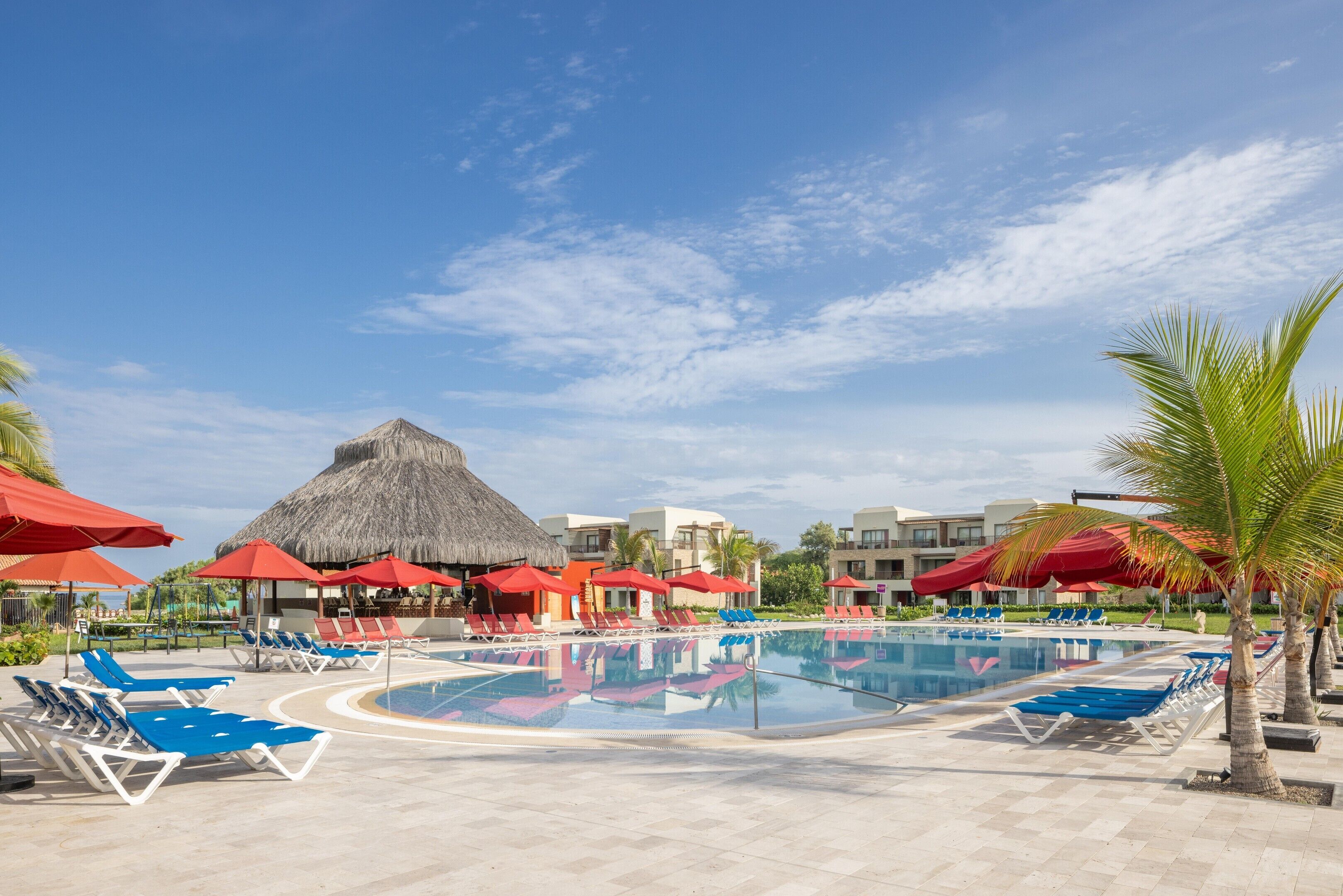 Un área de piscina con sillones de descanso y sombrillas, una estructura de techo de paja y edificios modernos al fondo.