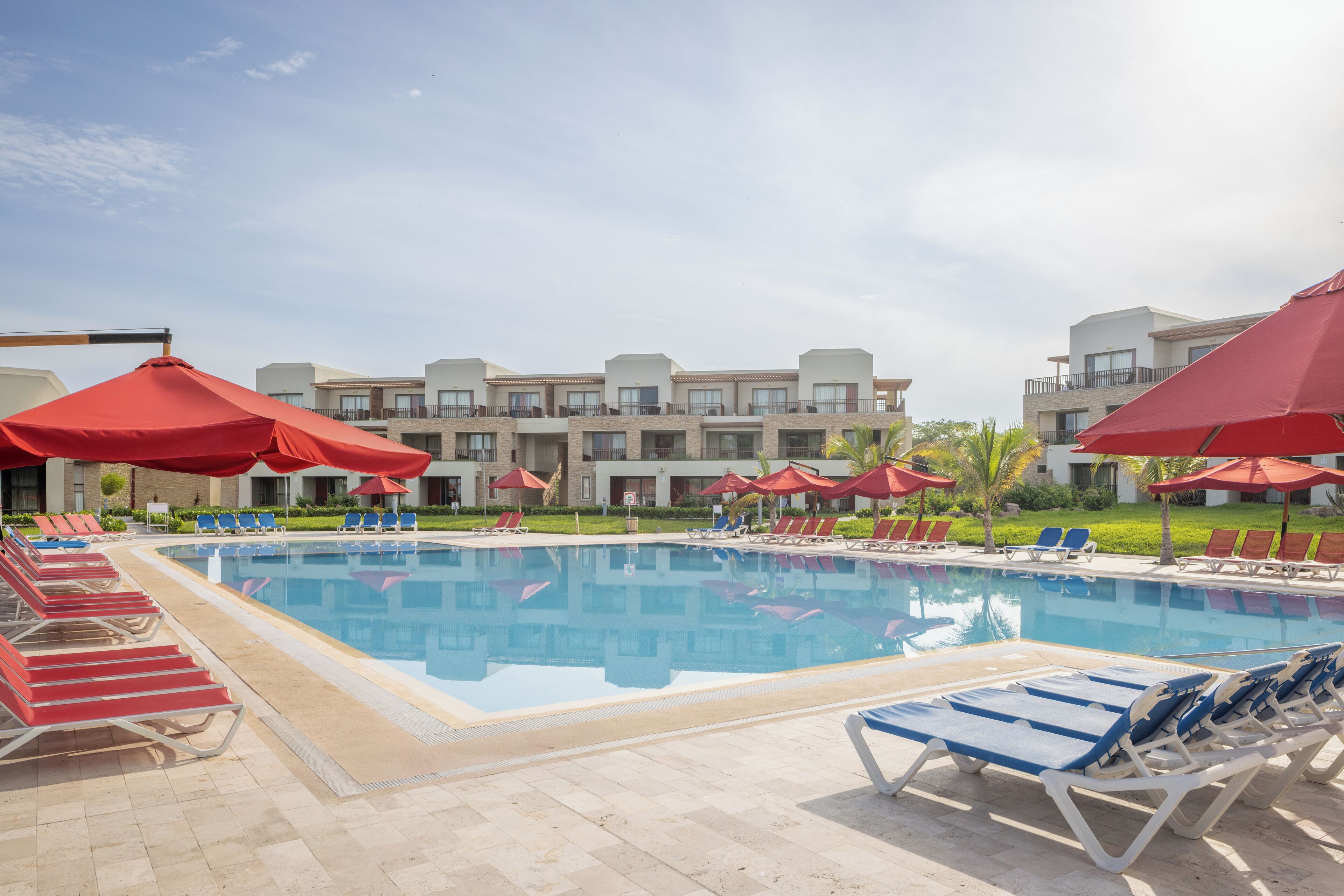 A swimming pool with red umbrellas and lounge chairs, surrounded by a building with balconies.