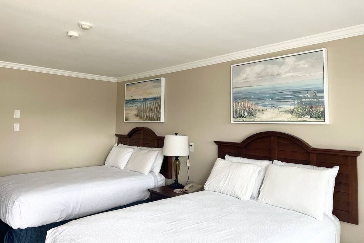 A hotel room with two beds, a nightstand with a lamp, and two paintings on the walls.