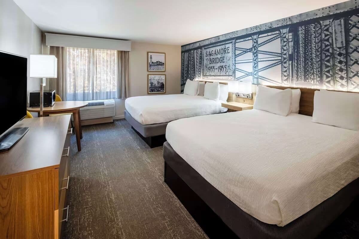 A hotel room with two beds, a desk with a lamp, a TV, and a large wall tapestry.