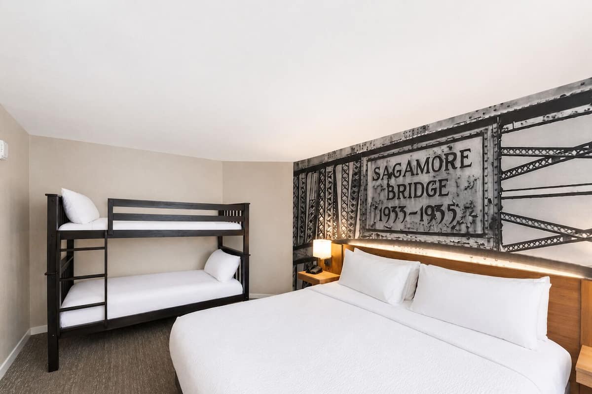 A hotel room with a bunk bed, a single bed, a nightstand with a lamp, and a wall with a historical plaque.