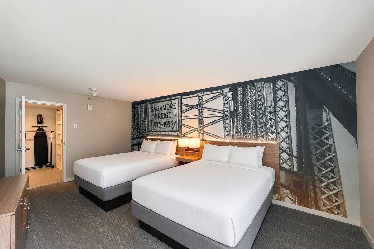 A hotel room with two beds, a wardrobe, a mirror, a wall with a bridge image, and a nightstand with a lamp.