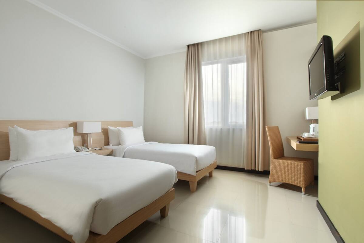 Habitación de hotel con dos camas individuales, un escritorio con silla, televisor montado en la pared y ventana con cortinas.