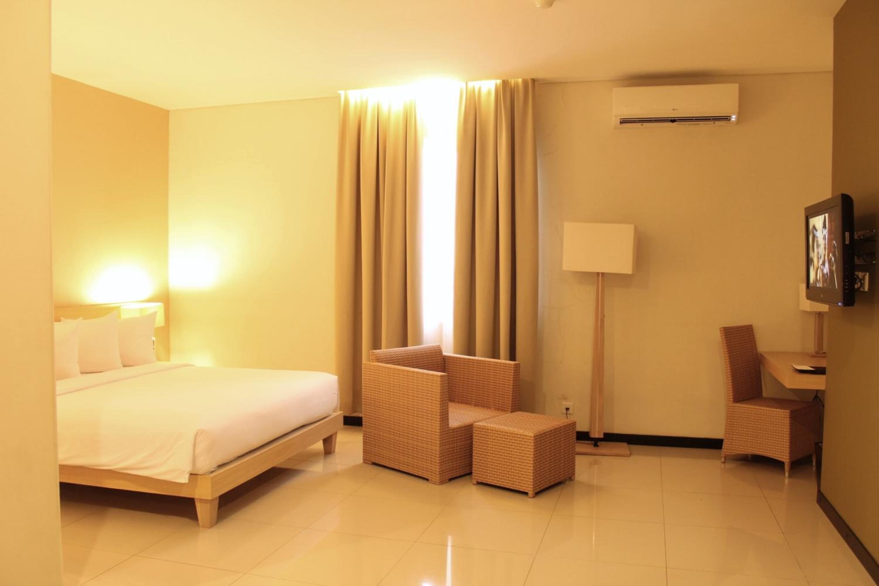 A hotel room with a bed, a seating area with a chair and a small table, a lamp, and an air conditioning unit.