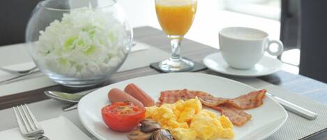 A breakfast plate with eggs, bacon, tomatoes, and mushrooms, a glass of orange juice, and a bowl of white flowers.