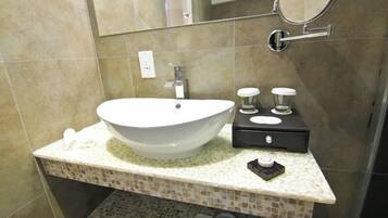Un baño con un lavamanos blanco tipo lavabo, un tocador negro pequeño con dos botellas, y un espejo sobre el lavamanos.