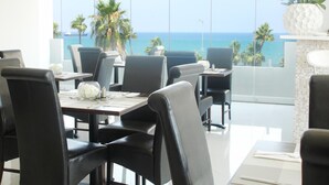 A dining area with a view of the ocean, featuring black chairs, white tables, and a white decorative vase. - Achilleos City Hotel (Larnaca)