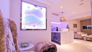 A modern living room with a large wall-mounted screen, a kitchen area, and a seating area with a sofa and a coffee table. - Achilleos City Hotel (Larnaca)