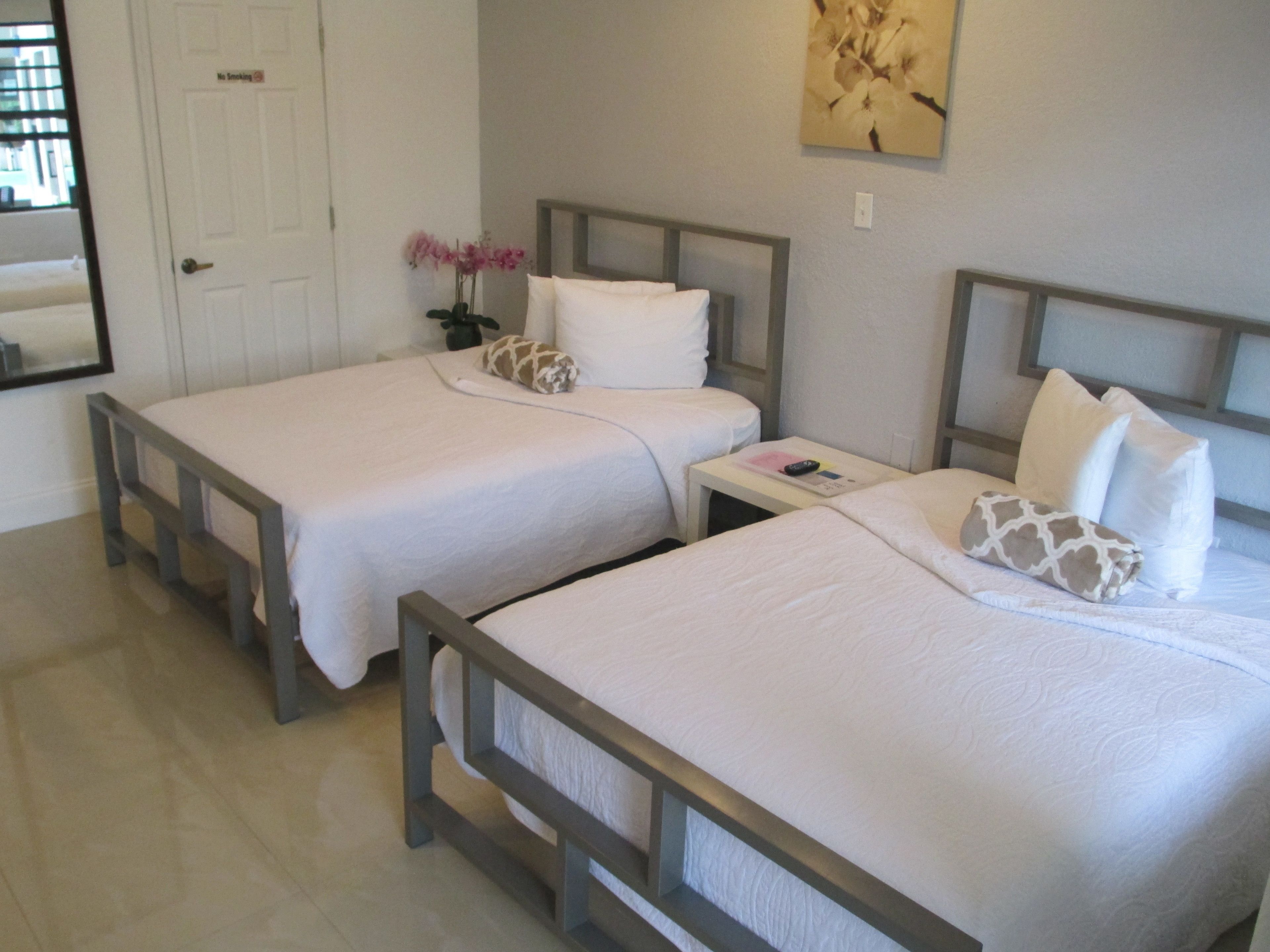 A hotel room with two single beds, a nightstand with a remote control, a small potted plant, and a painting on the wall.