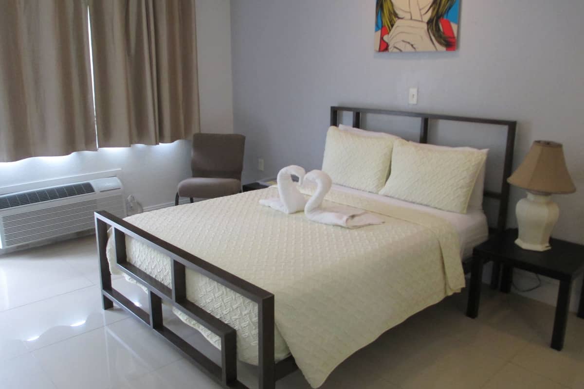 A hotel room with a bed, a chair, a wall-mounted painting, a lamp, and an air conditioning unit.