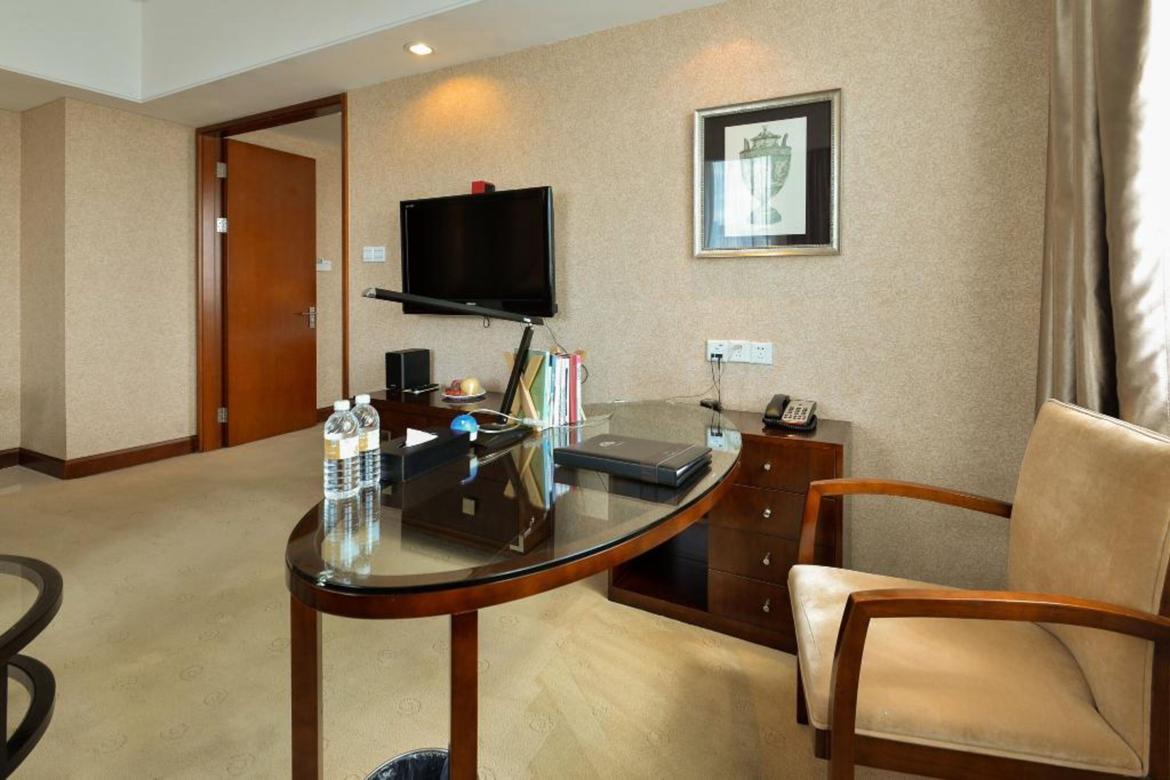 A hotel room with a round glass table, a flat-screen TV mounted on the wall, a framed picture, and a wooden chair.