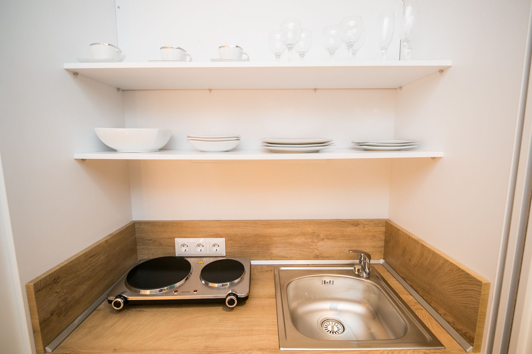 Une petite cuisine équipée d’un évier, de deux bouilloires électriques, d’une petite cuisinière et d’étagères où sont rangés des bols, des assiettes et des verres.