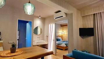 A hotel room with a wooden dining table, a small potted plant, a blue sofa, a bed with a blue blanket, a mounted TV, and a bathroom with a mirror and sink.