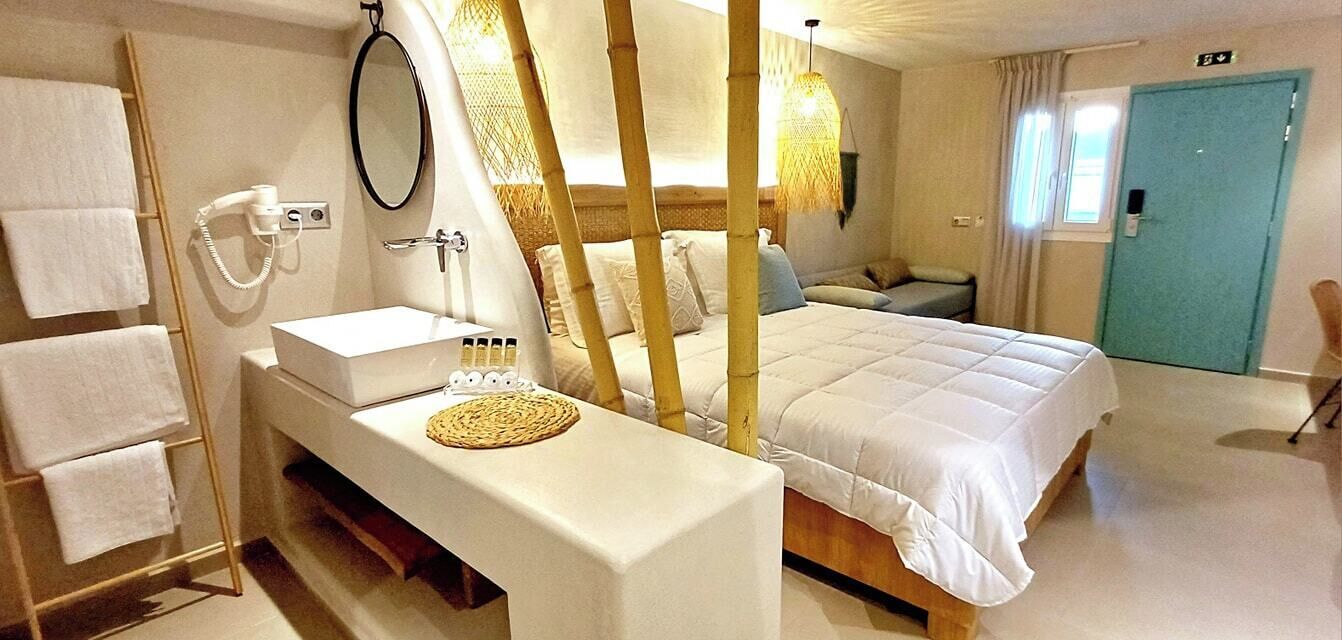 A hotel room with a bed, a bedside table with a mirror, a sink, and a towel rack.